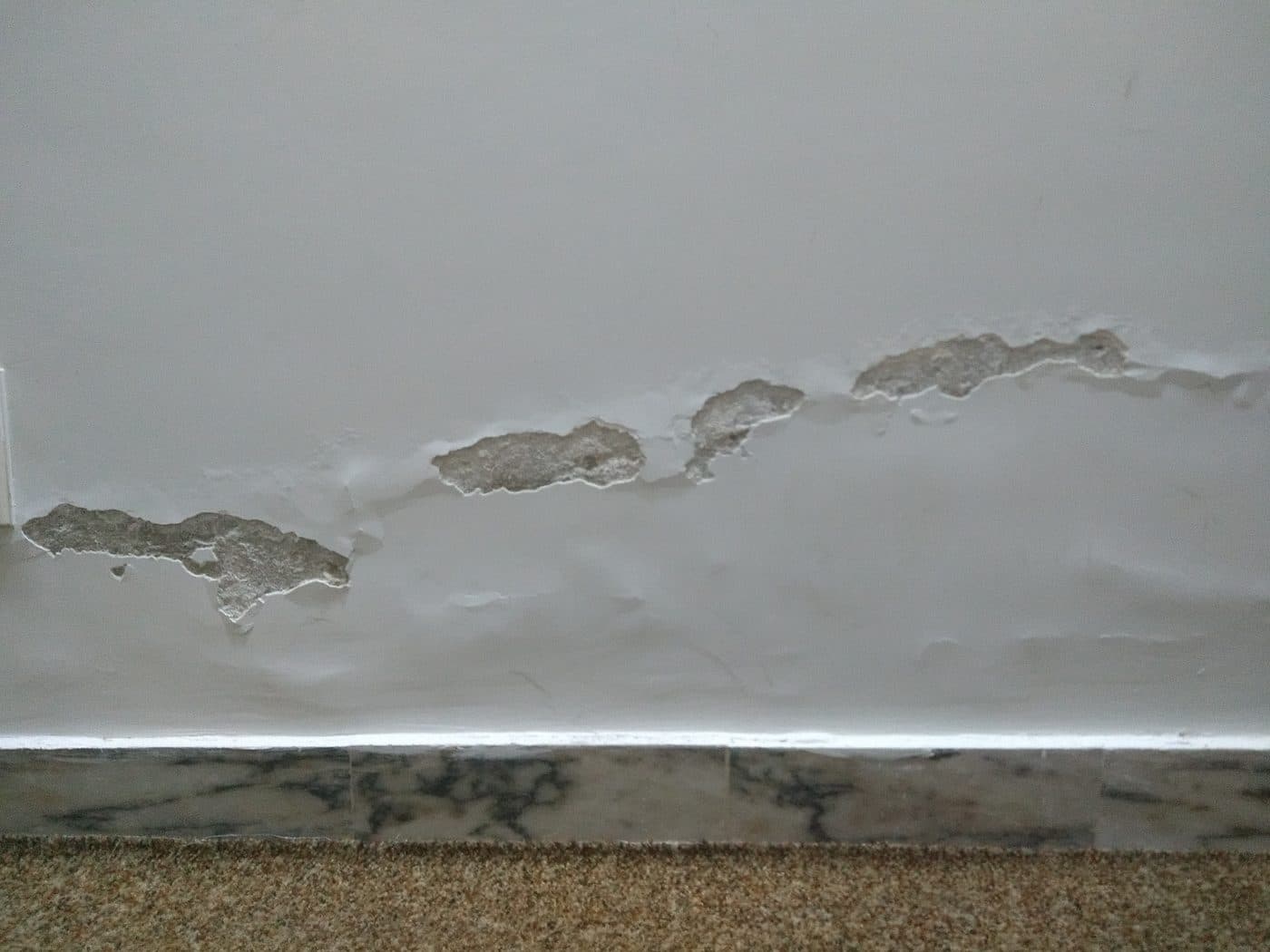 Parede interior com a tinta e o reboco a levantar numa faixa horizontal logo acima do rodapé